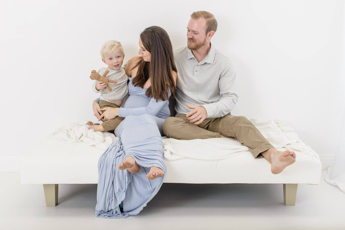 A family of three sits on a white couch in a bright room. The mother, in a light blue dress, looks at the young boy holding a small stuffed animal. The father, wearing a light shirt and khaki pants, smiles at them both.