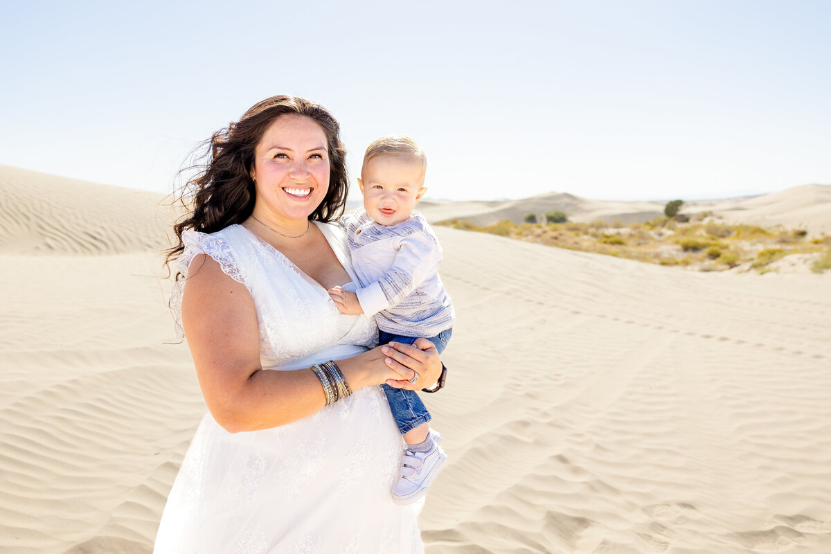 A mother in a flowing white dress holds her young son in the middle of vast desert sand dunes. The wind plays with her long dark hair as both smile brightly under the warm sun.