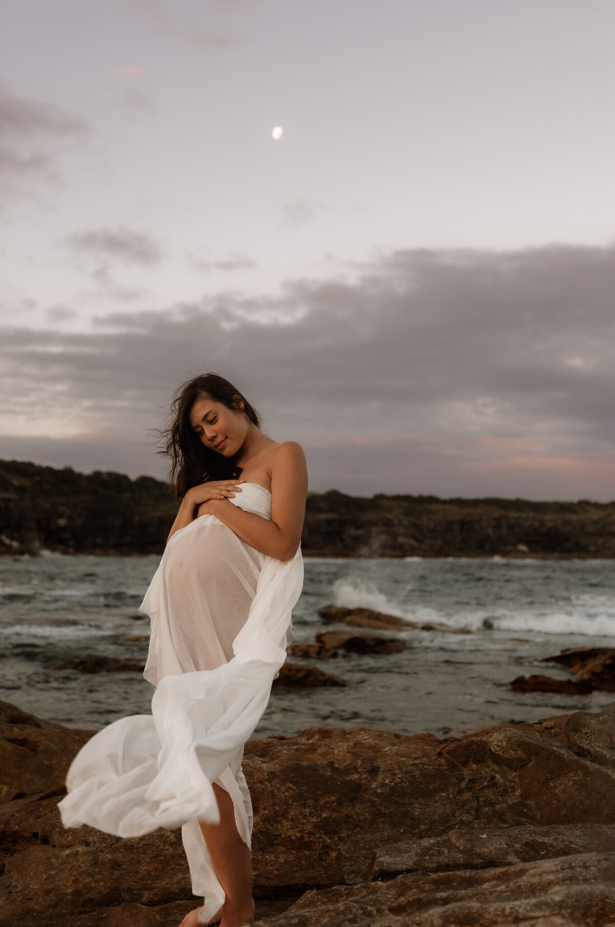 Pregnant woman wrapped in a flowing white fabric standing on coastal rocks at sunset in Sydney, with soft moonlight and a gentle breeze creating an ethereal maternity portrait.