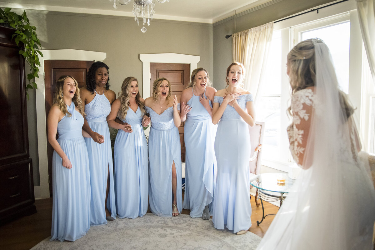 A group of bridesmaids in light blue dresses react to seeing the bride for the first time on her wedding day.
