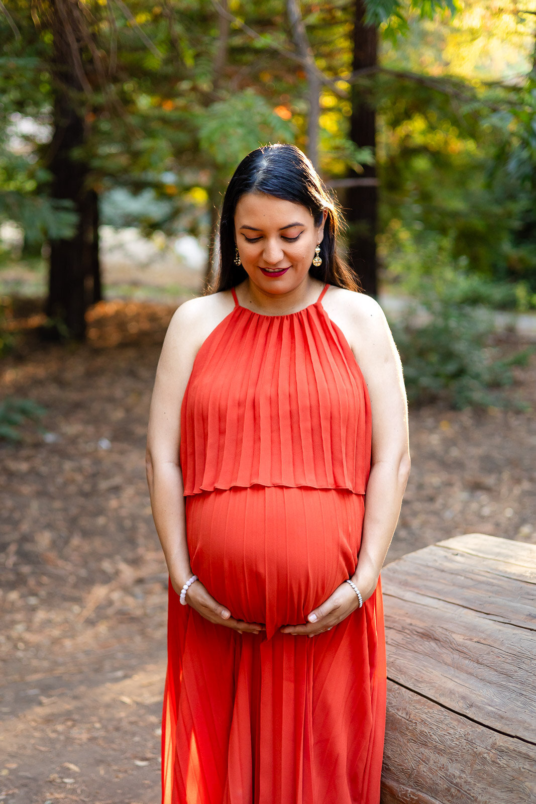 Pregnant woman looking down at her hands on her baby bump in a pleated orange dress – Ellobelle Photography