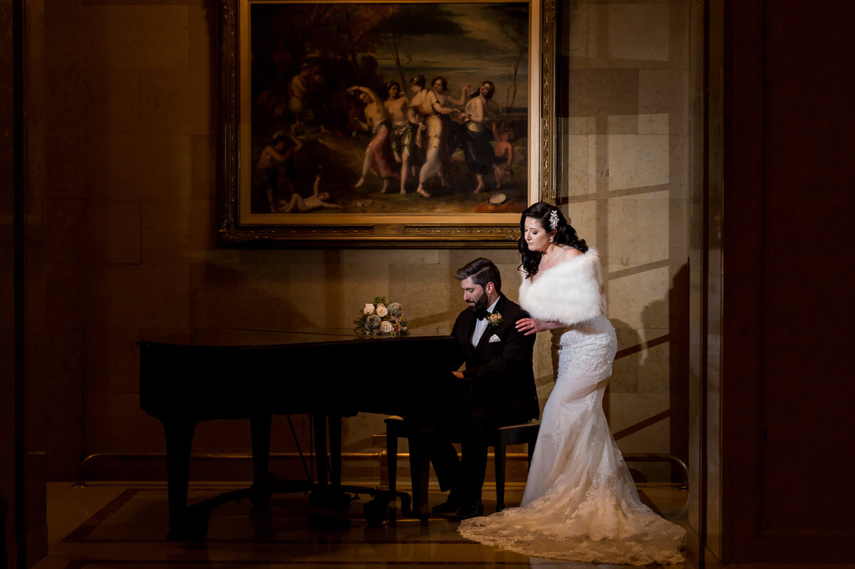 a dramatic photo of a groom in a tuxedo playing a grand piano while his elegant bride leans over and watches.  Captured by Ottawa wedding photographer JEMMAN Photography