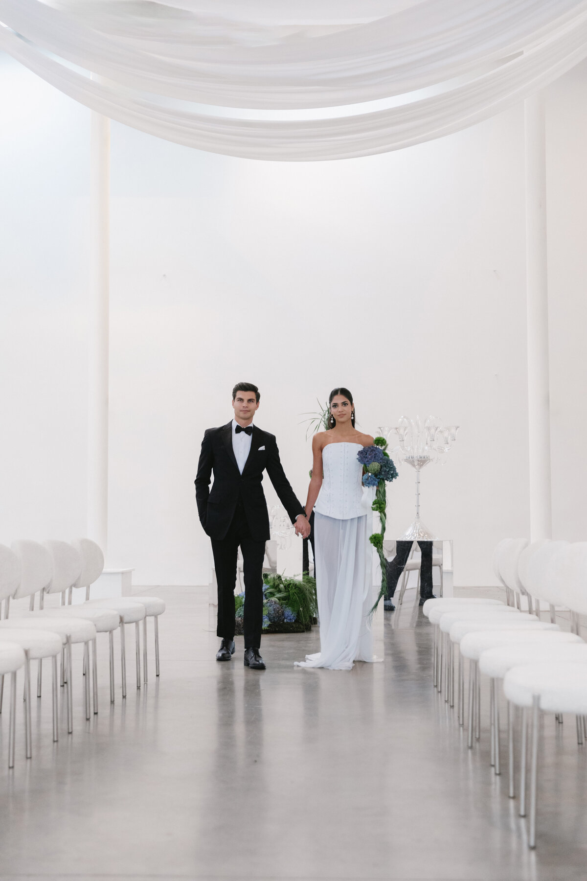Fashion-forward bride and groom walking together in a minimalist white studio aisle decorated with sculptural florals in NYC.