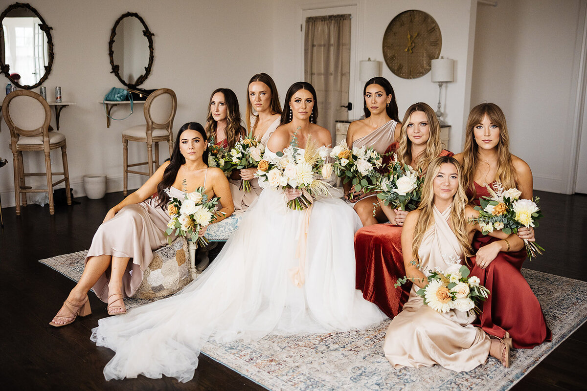 Bride poses with bridesmaids in rust, red, and champagne bridesmaid dresses.