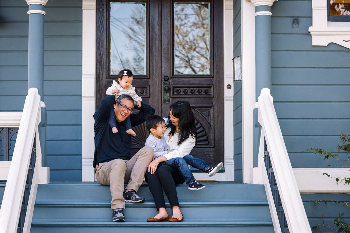 mom and dad sitting on front porch of blue house in Alameda with son and toddler daughter interacting with each other and laughing