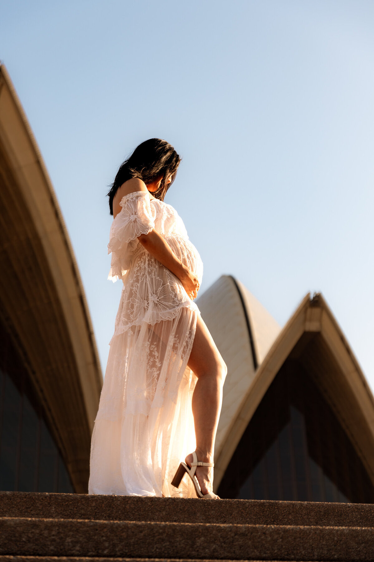 Pregnant woman in a sheer white dress cradling her belly in front of the Sydney Opera House sails during a sunrise maternity session
