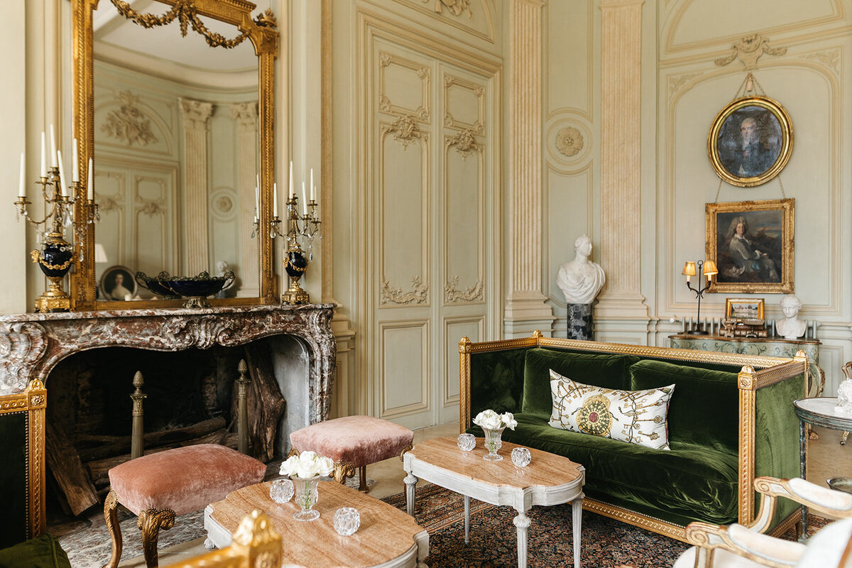 A luxurious French-style sitting room featuring a marble fireplace, gold accents, green velvet furniture, and classical artwork.