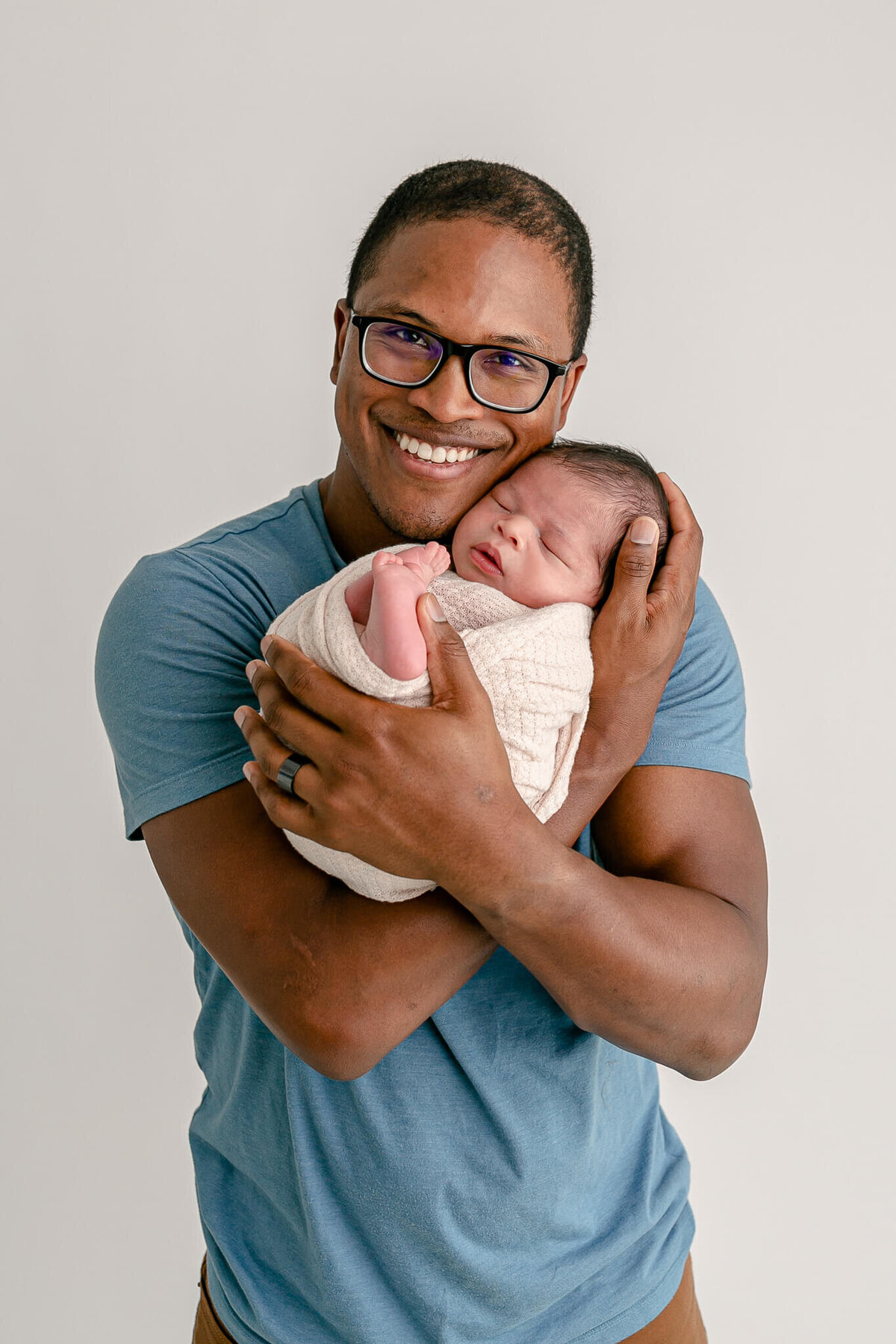 Dad with glasses wearing a blue t-shirt is holding baby in his arms with baby up close to his cheek. Baby is sleeping and wrapped in a beige wrap with his little toes peeking out. Dad is smiling proudly at the camera.