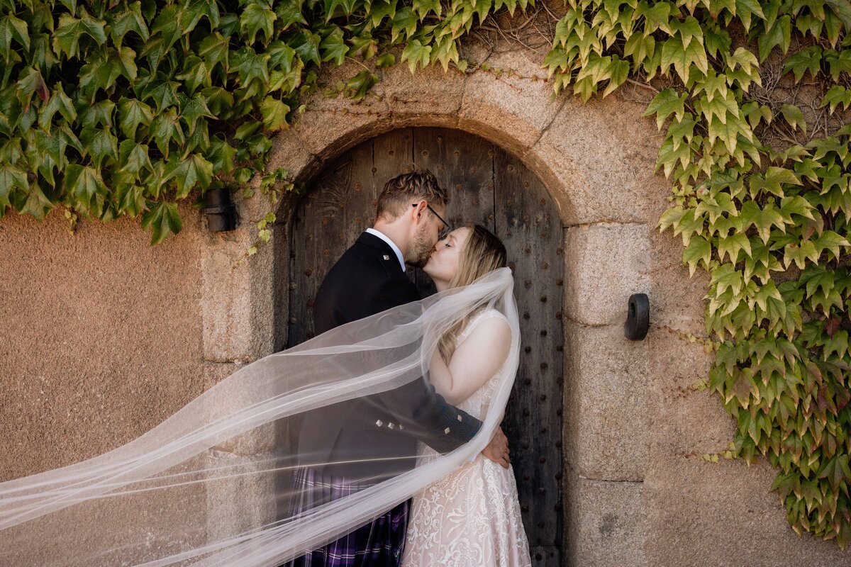 A brides veil blows in the wind as she's photographed kissing her groom,