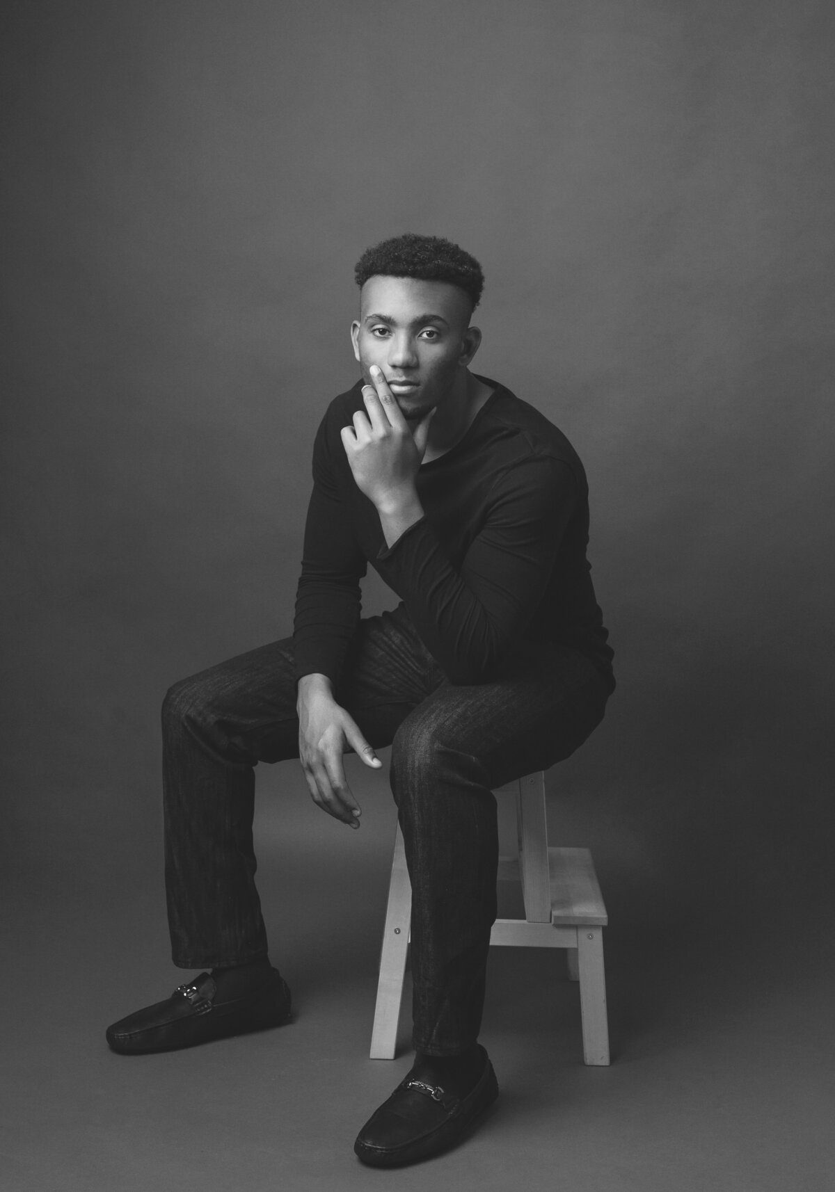 Professional portrait of young man sitting on a stool, resting head on his left hand.