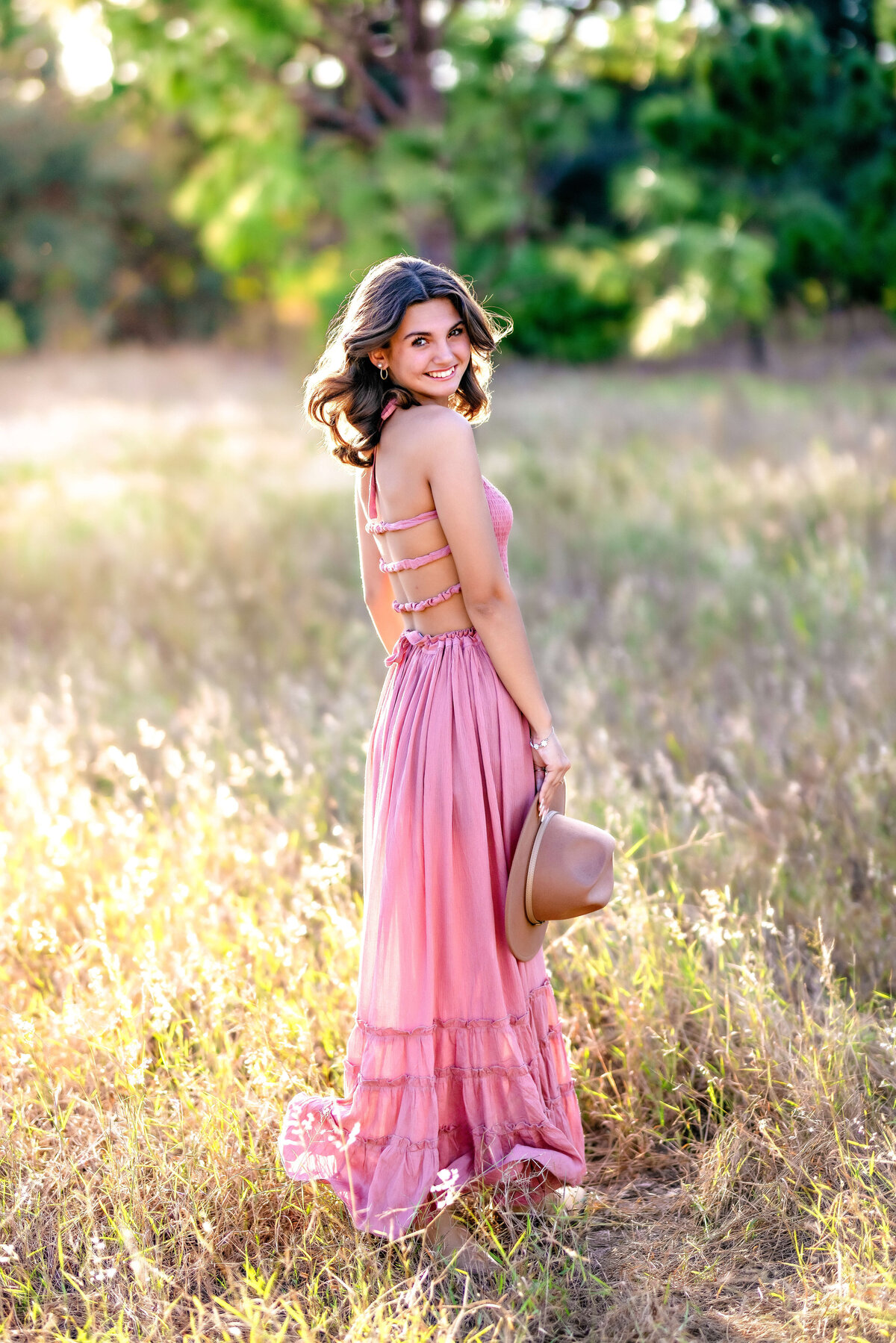 High school senior in a pink dress standing still in tall grass with soft sunset light and cheeky smile