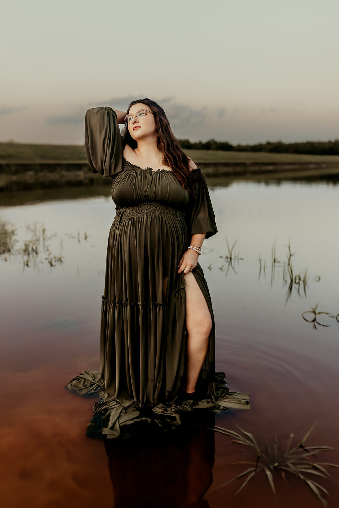 A person in a flowing dark green dress stands in shallow water, in a nature setting with a calm lake and grassy shoreline at dusk.