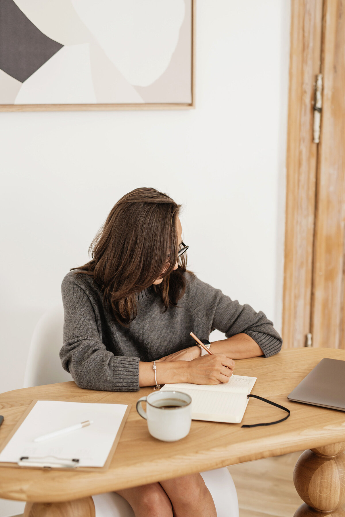 Close-up of a woman in a gray sweater writing in a notebook at a wooden desk, with a laptop partially visible on the right. This image introduces Fractional COO services, supporting streamlined systems and backend clarity for creative firms without micromanagement.