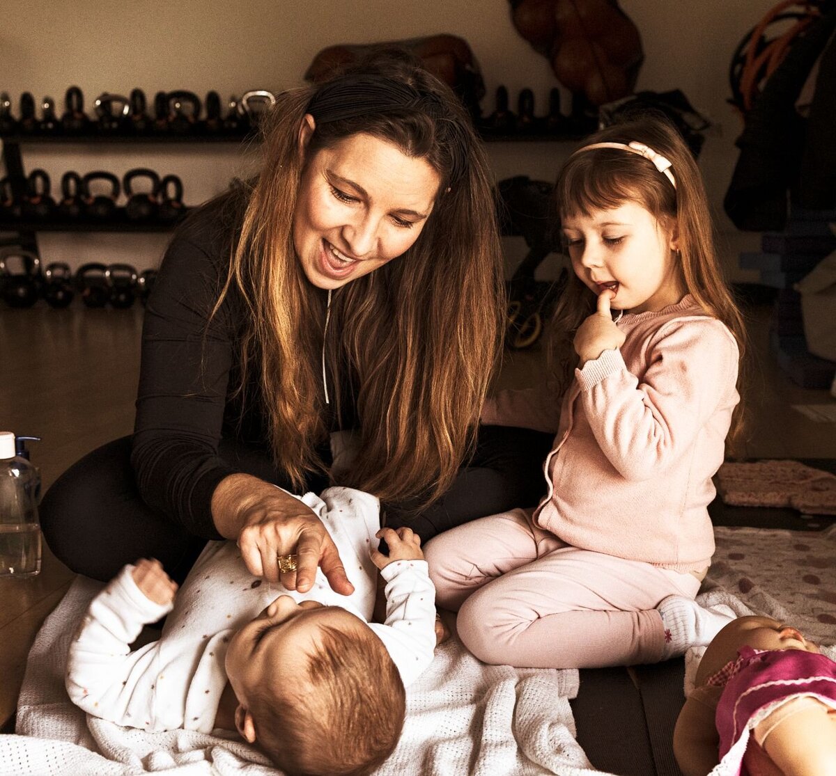 A heartwarming scene of a mother and her two young children on the floor: the baby looking up, the toddler observing thoughtfully, and a doll resting close