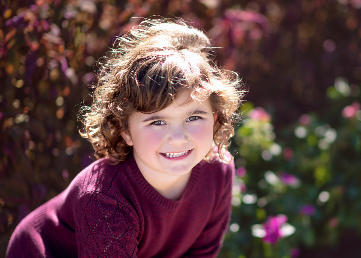 Child with curly hair in a purple sweater with a floral background smiles for her Iowa Milestone Photos.