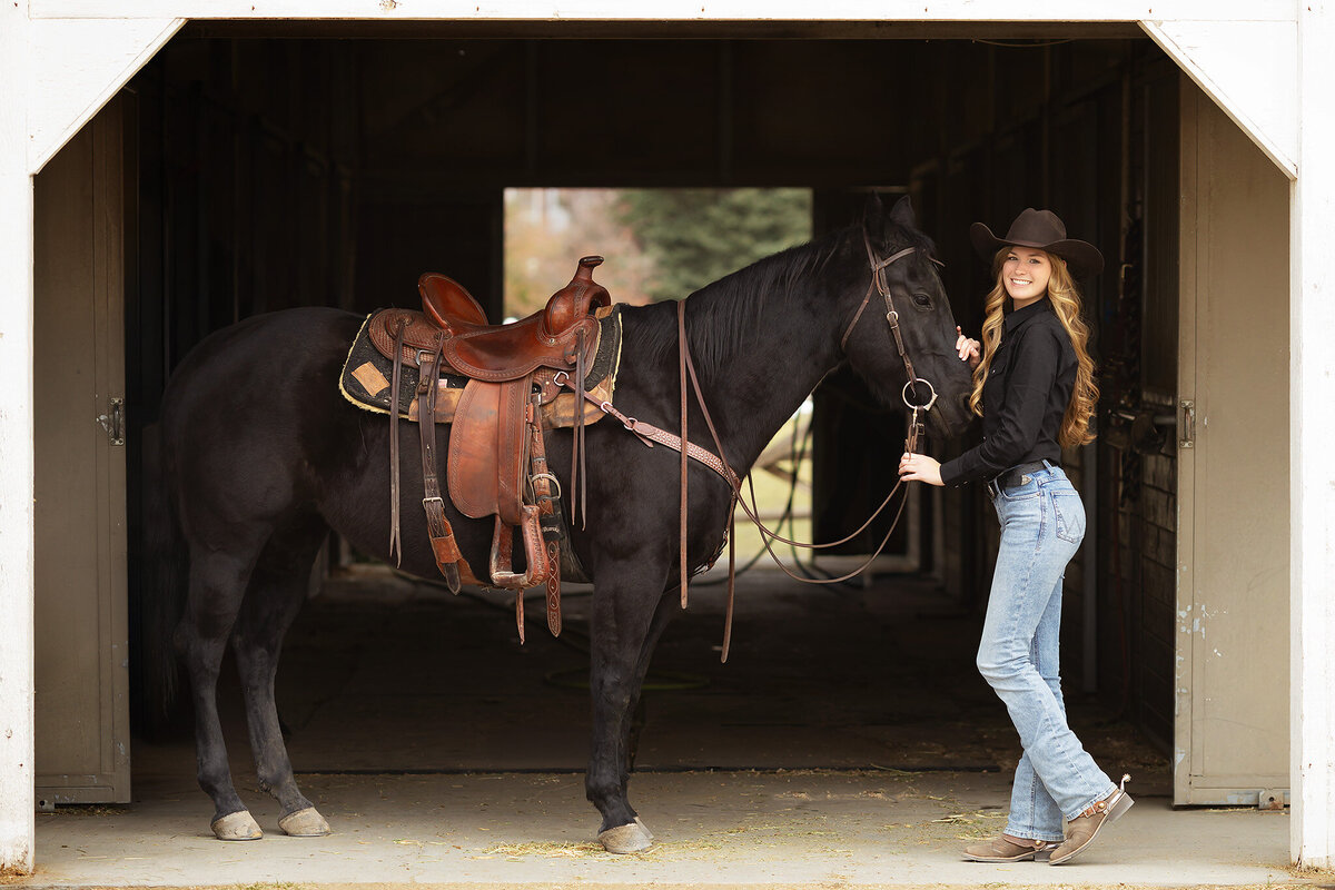 A senior girl in jeans and a black shirt stands with her horse in a barn, captured by a Dallas senior photographer.