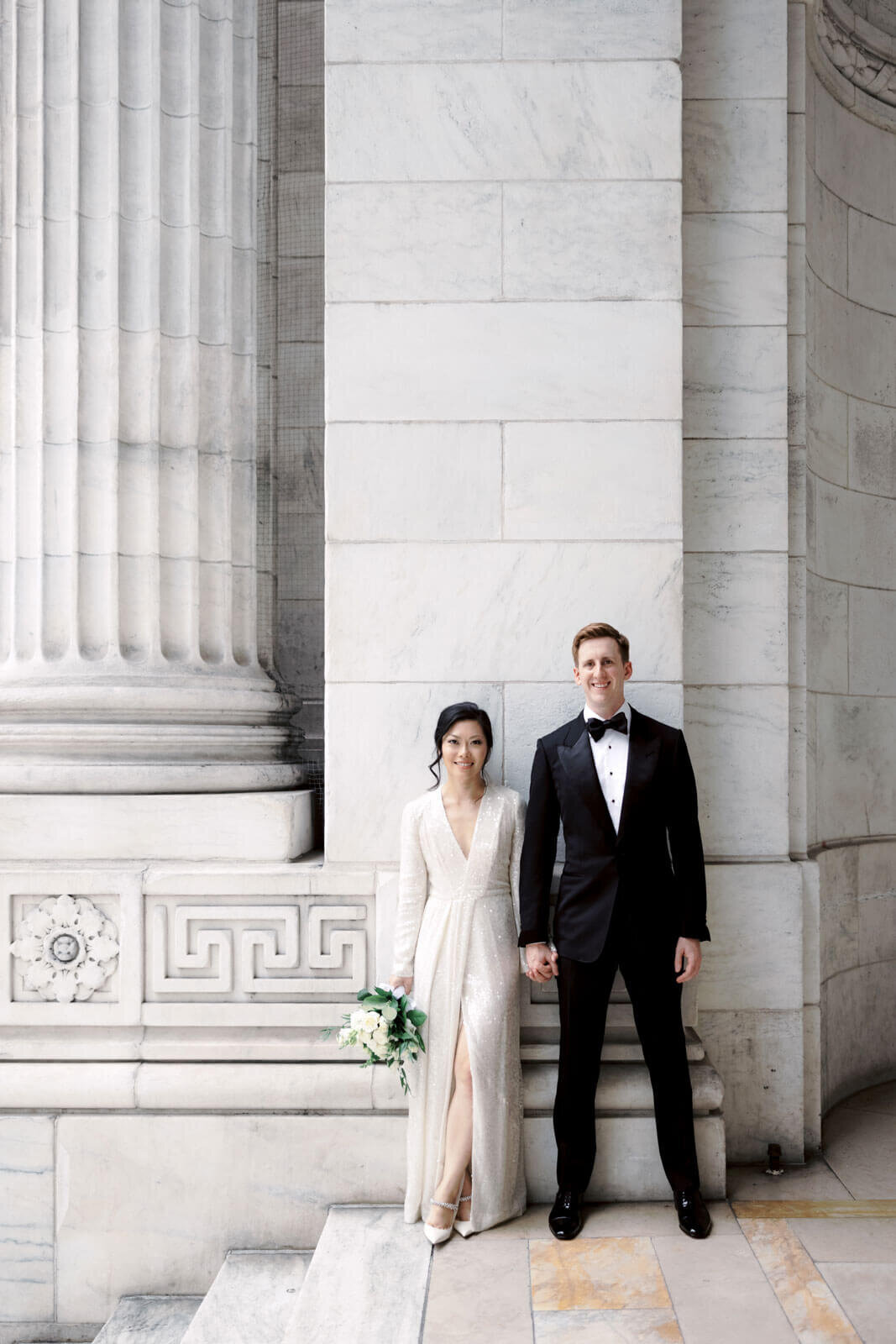 The bride and the groom are on top of the New York Public Library's grand staircase with large marble columns and walls.