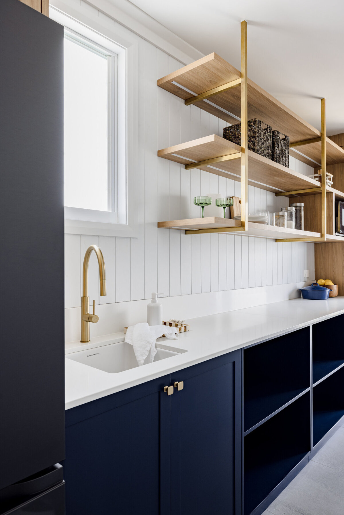 Modern kitchen in a luxury home design, featuring navy cabinets, a white countertop, a gold faucet, and open wooden shelves adorned with decorative items. A prime example of bespoke interior design in Newcastle NSW.