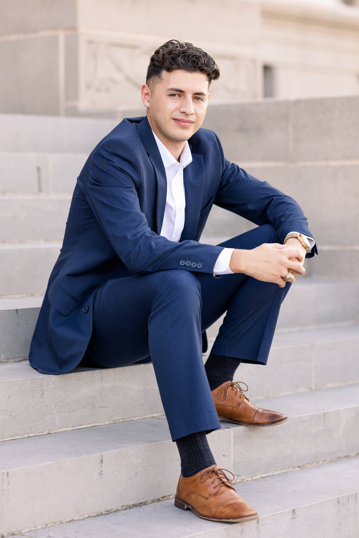 A polished senior portrait of a young man in a navy blue suit, seated on stone steps in College Station, Texas. A confident and timeless image perfect for graduation announcements, photographed by Jennifer Lynn Photography.