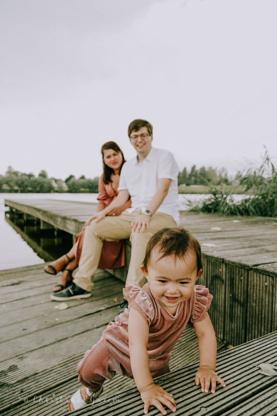 Toddler crawling on wooden pier – A cheerful baby girl in a dusty rose outfit crawls toward the camera while her parents sit smiling in the background near a peaceful lake.
