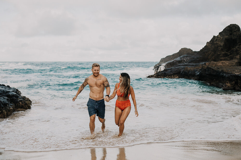 A couple running on the beach  with the waves crashing behind them.
