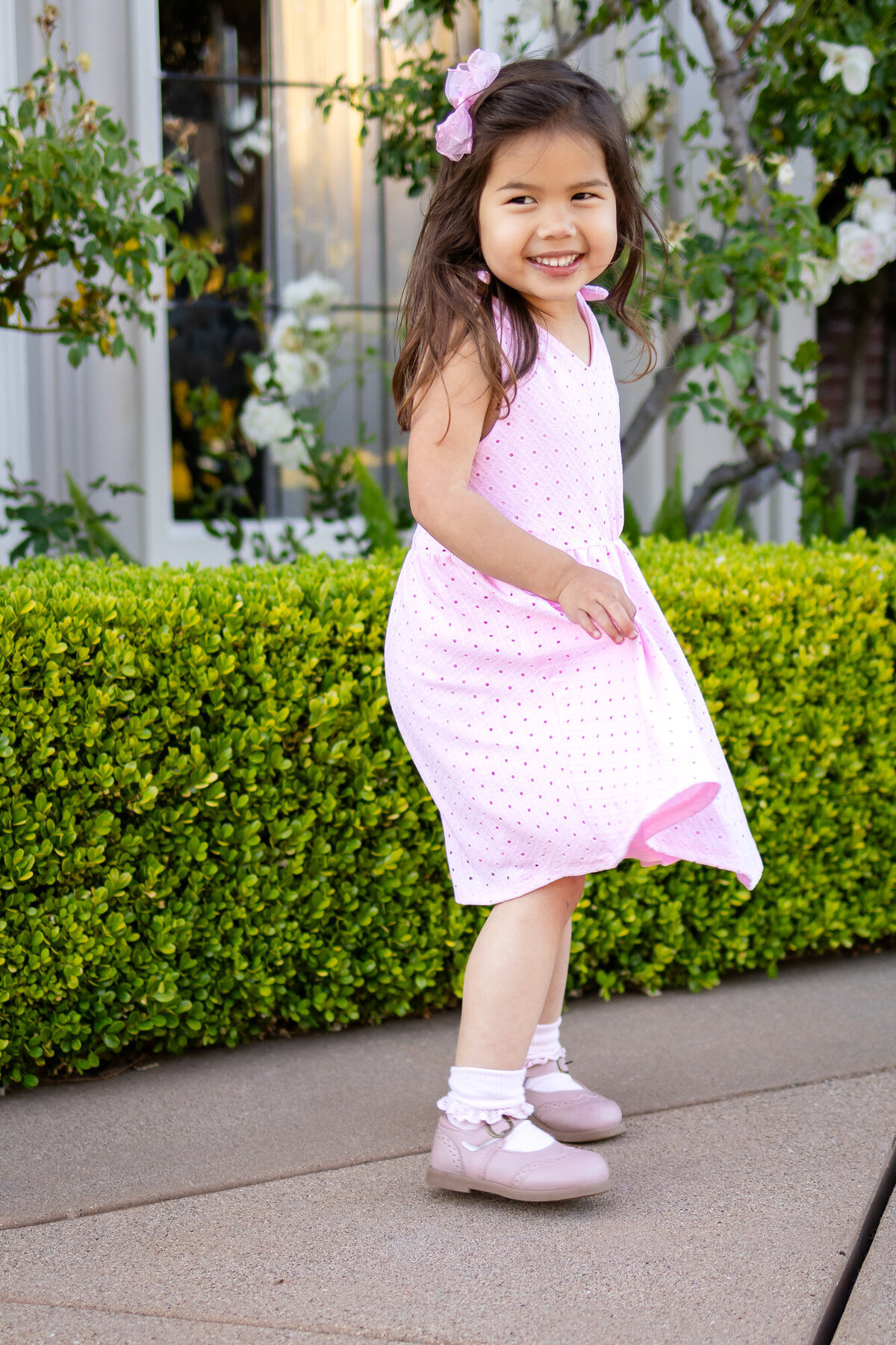 Bay Area Family Portfolio - Ellobelle Photography captures a young girl twirling joyfully in a pink eyelet dress, smiling brightly in front of green boxwood hedges.