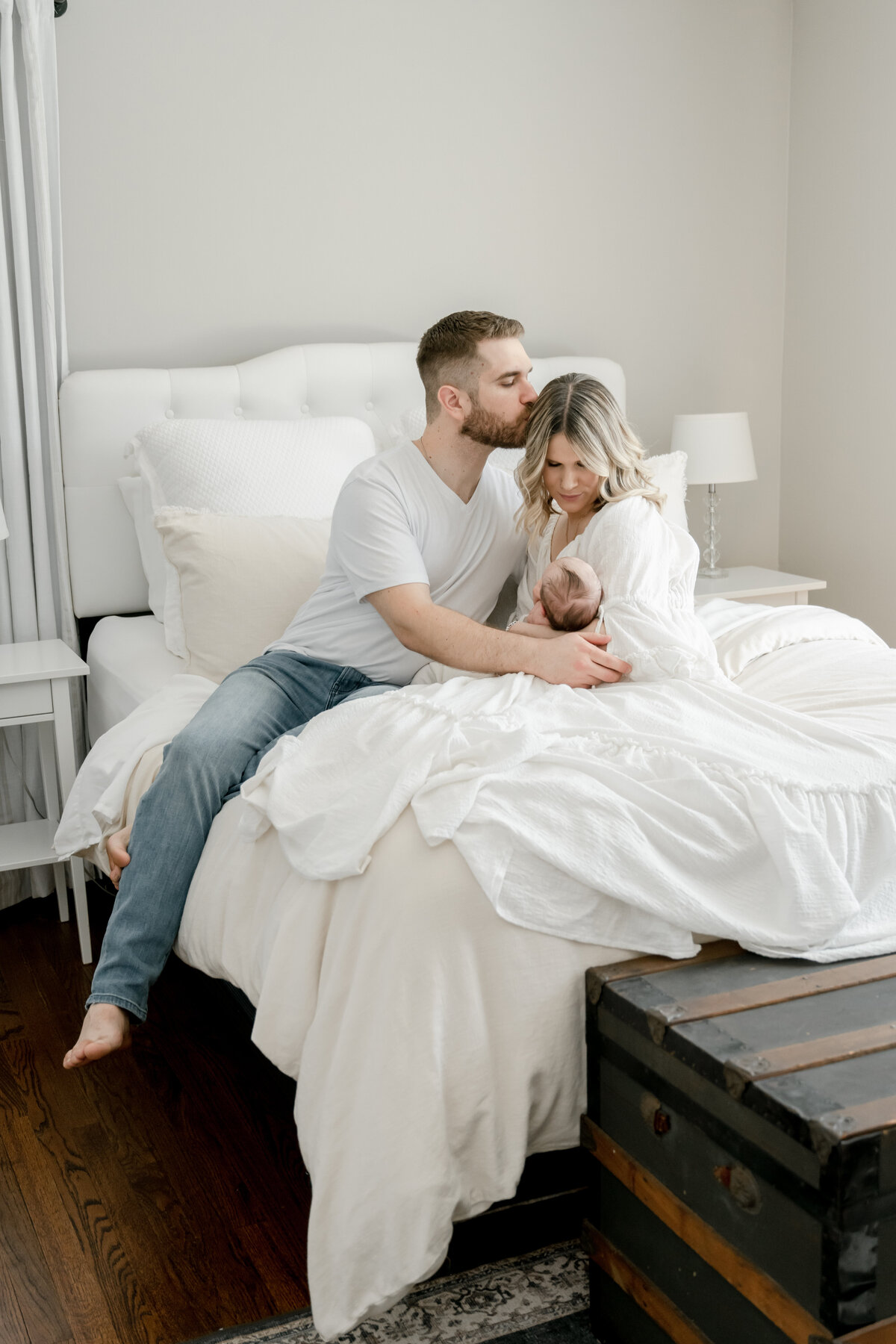 Dad is kissing the mum who is holding their newborn baby in her arms looking down. They are sitting on a bed with a white cover during their in home newborn session in New Jersey