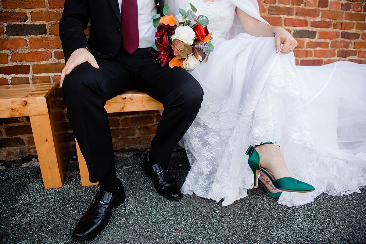 Detail of the bride and groom's shoes. The bride is wearing emerald green heels with an ankle strap. The groom is wearing shiny black shoes. The groom is wearing a black suit with a white shirt and a maroon skinny tie. Th e bride has a flowing white  wedding dress with lace detailing an da small bouquet of yellow, orange, ivory and dark red flowers.