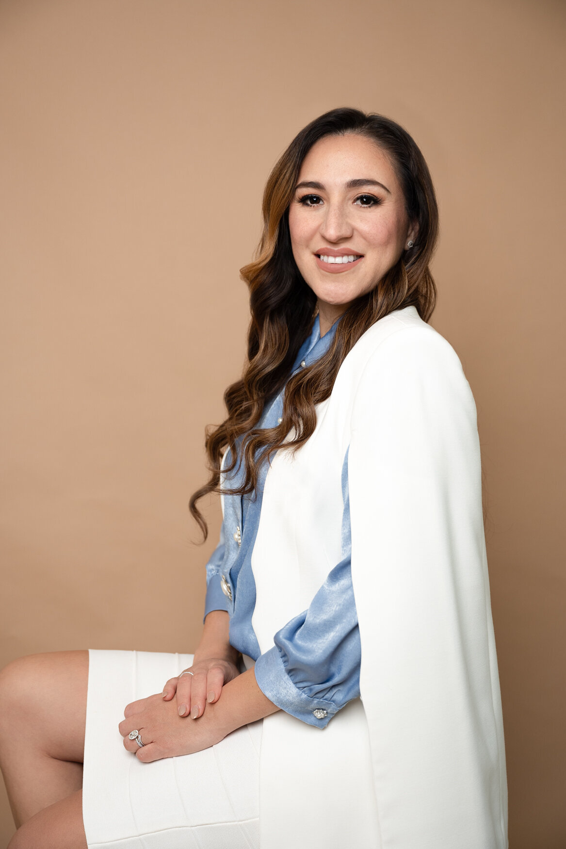 Professional portrait of a confident woman seated against a neutral beige backdrop, wearing a white blazer and a light blue satin blouse, radiating poise and approachability