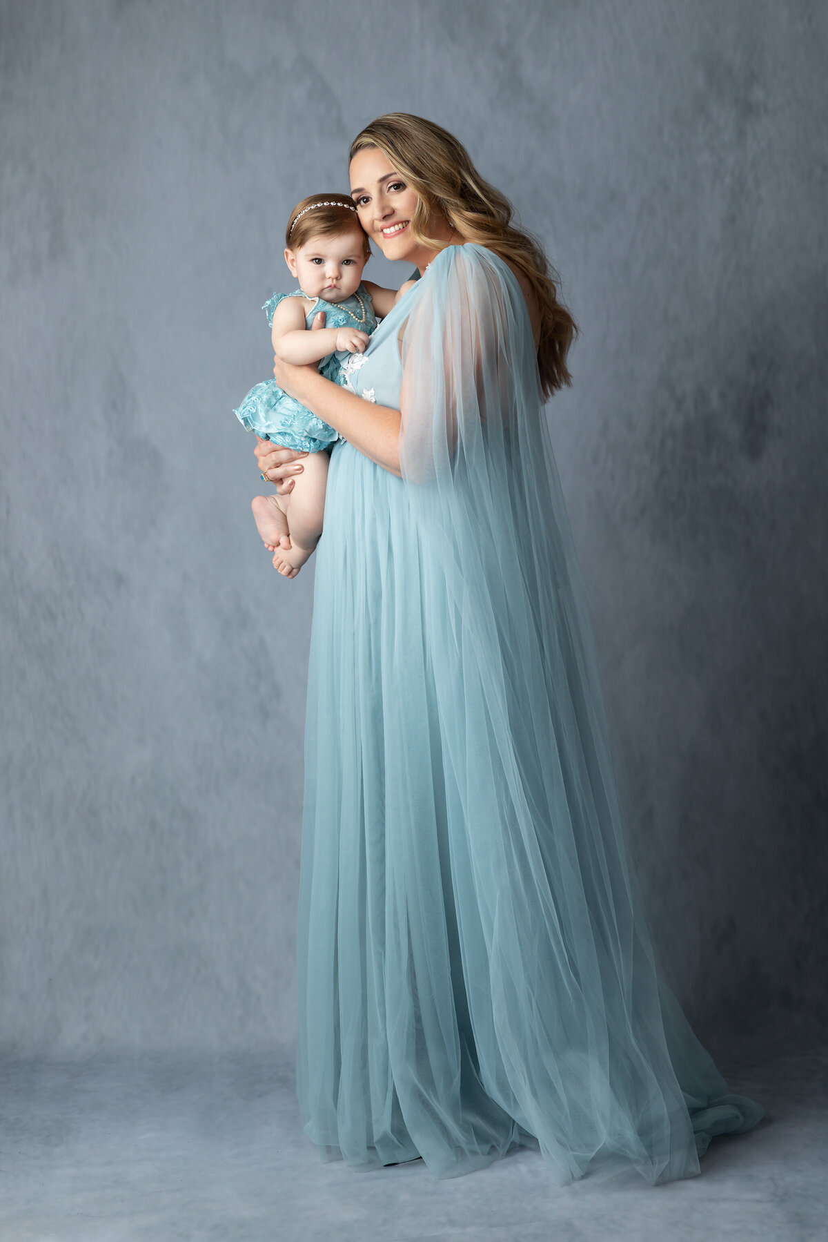 Full-length studio portrait of a mother in a flowing blue gown holding her baby girl, captured by a DFW milestone photographer.