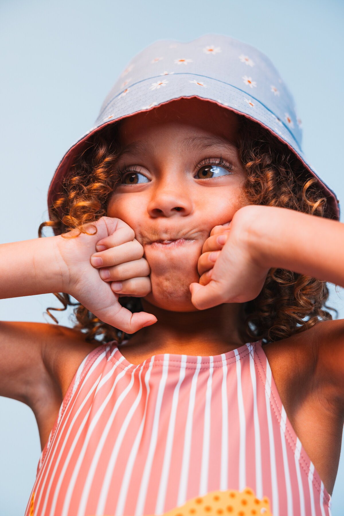 Playful child in a striped Swim Essentials swimsuit making a funny face, wearing a light blue daisy-print bucket hat against a soft blue background.