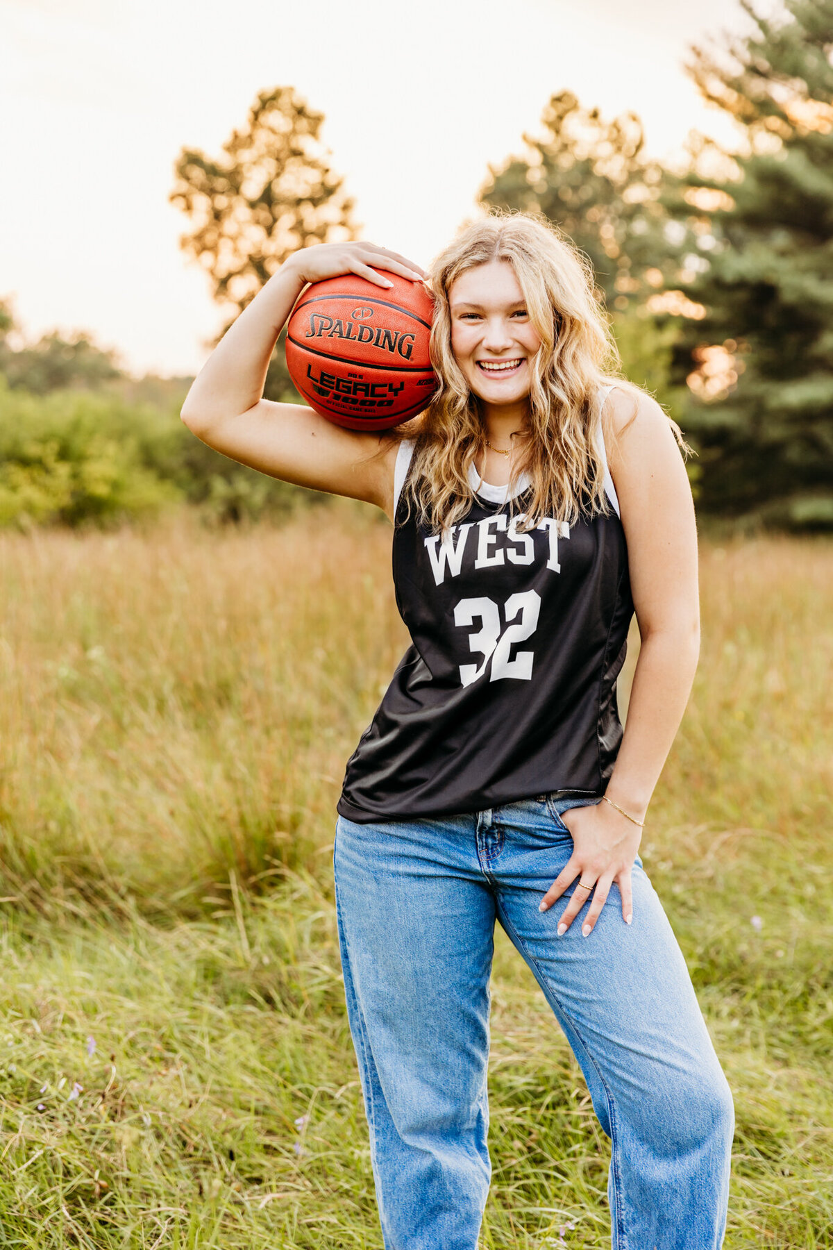 gorgeous blonde teenage girl in an Oshkosh West High School basketball jersey holding a basketball and laughing for her senior photo session.