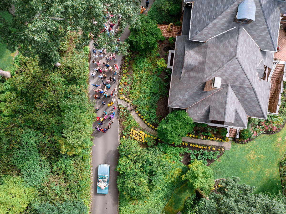 Aerial view of a residential street with a group of cyclists riding beside a vintage car. Lush green trees and bushes surround a large house with a gray roof. The path curves through the landscape, creating a scenic suburban setting.