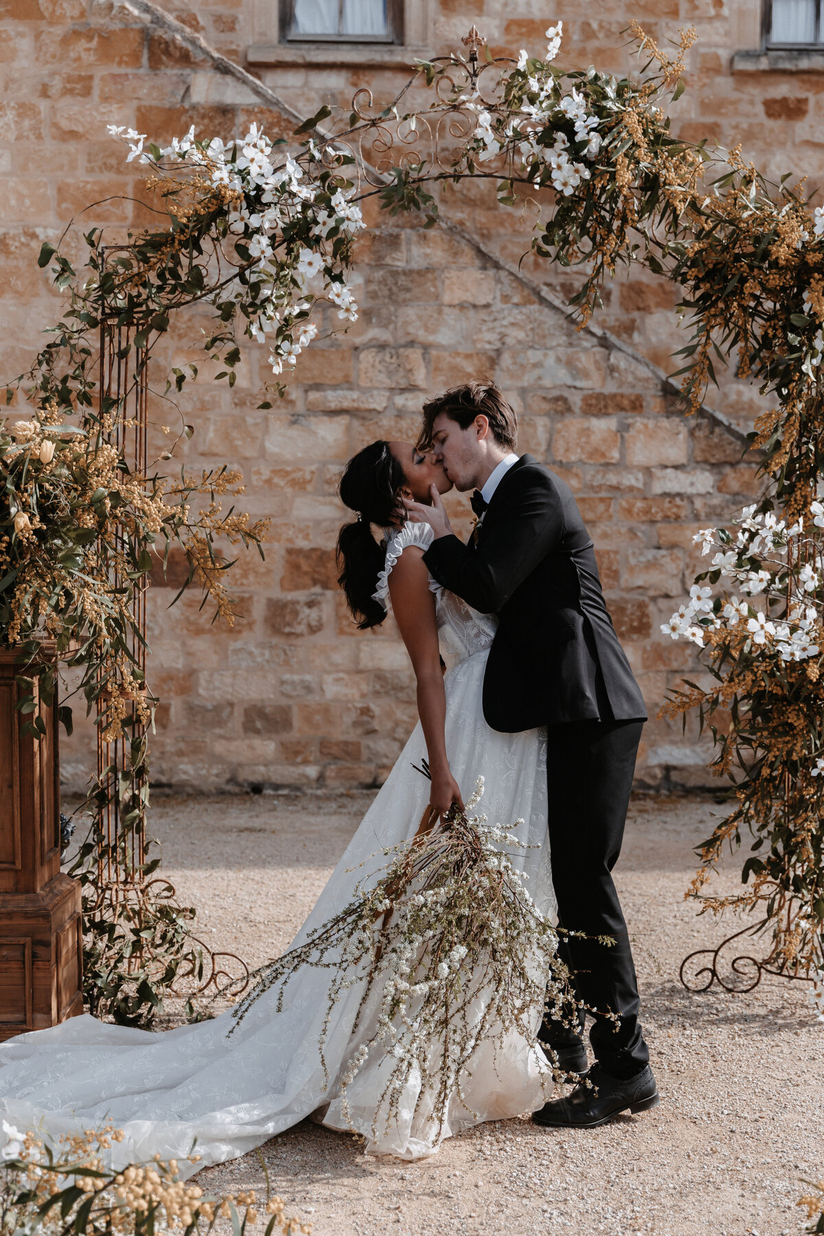 Bride and groom share a romantic kiss under a floral arch at Sunstone Winery, a rustic vineyard wedding venue.