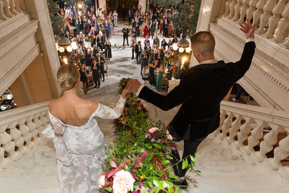 A bride and groom are standing on a balcony holding hands