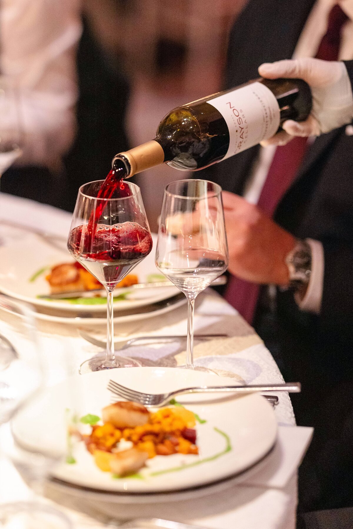 A person in formal attire pours red wine from a bottle into a glass at a table set with plates of gourmet food and empty wine glasses, creating an elegant dining atmosphere.