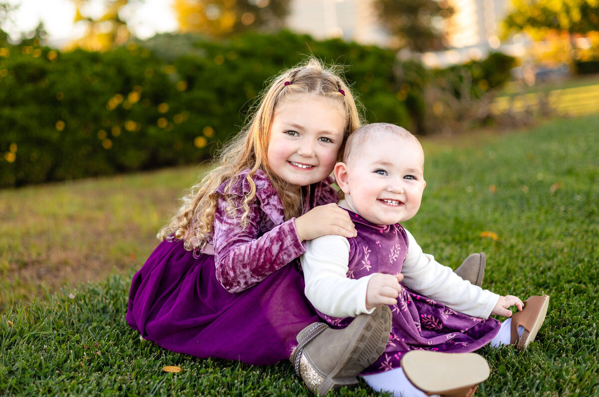 Sisters in matching purple dresses laughing on green grass at a San Jose park – Bay Area Family Portfolio – Ellobelle Photography