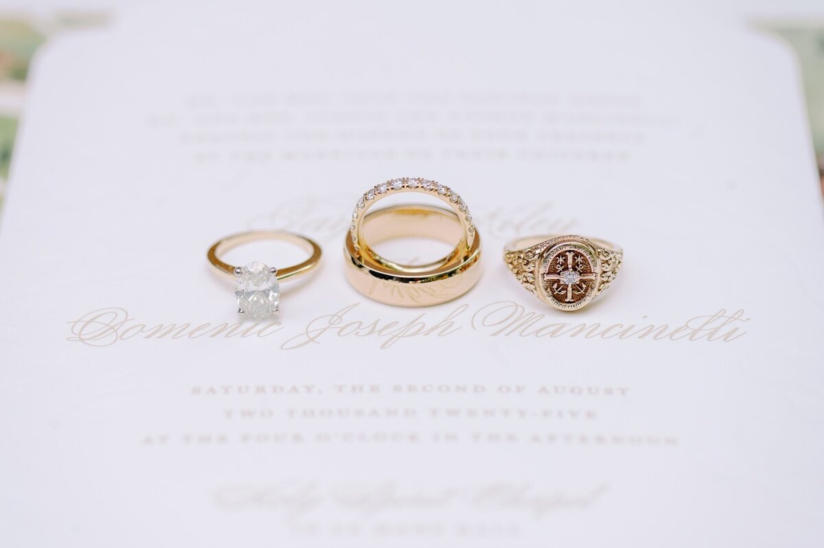 an engagement ring, wedding rings, and a class ring sitting on top of a wedding invitation