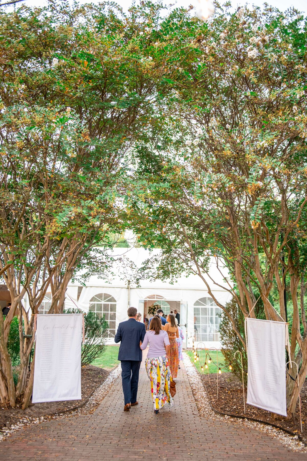 Three people walk down a brick path under an archway of trees towards a white wedding tent. Two large seating charts are displayed at the entrance, surrounded by greenery and soft lighting.