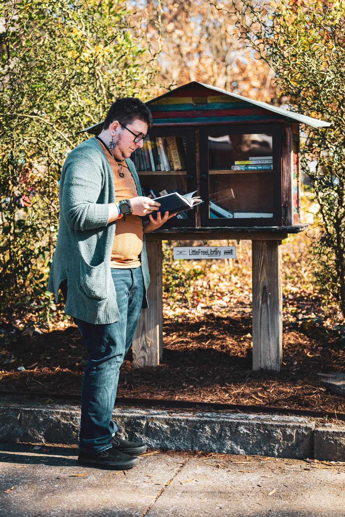 A person standing in front of a free library reading a book.