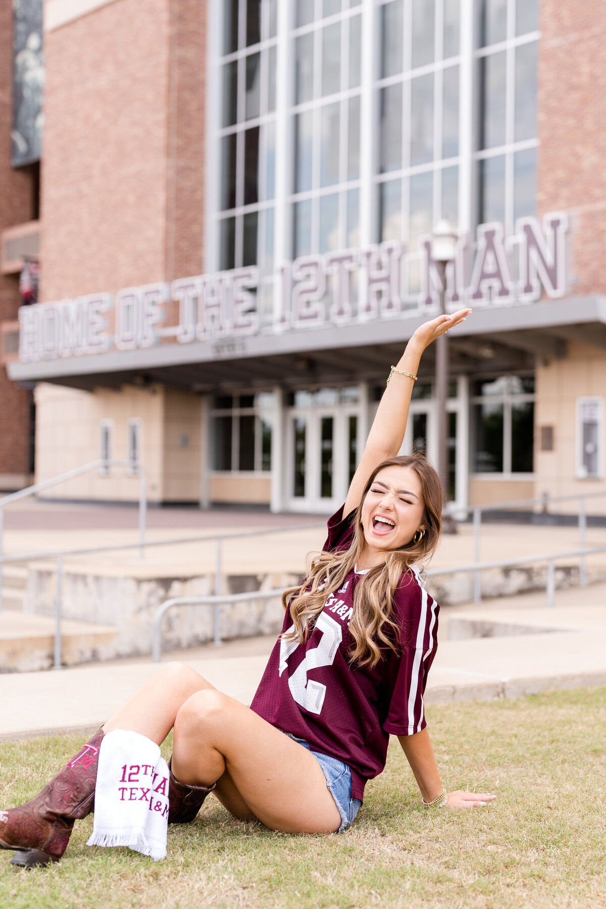 Texas A&M senior girl sitting down and celebrating in front of Kyle Field while wearing maroon Aggie jersey and 12th man towel