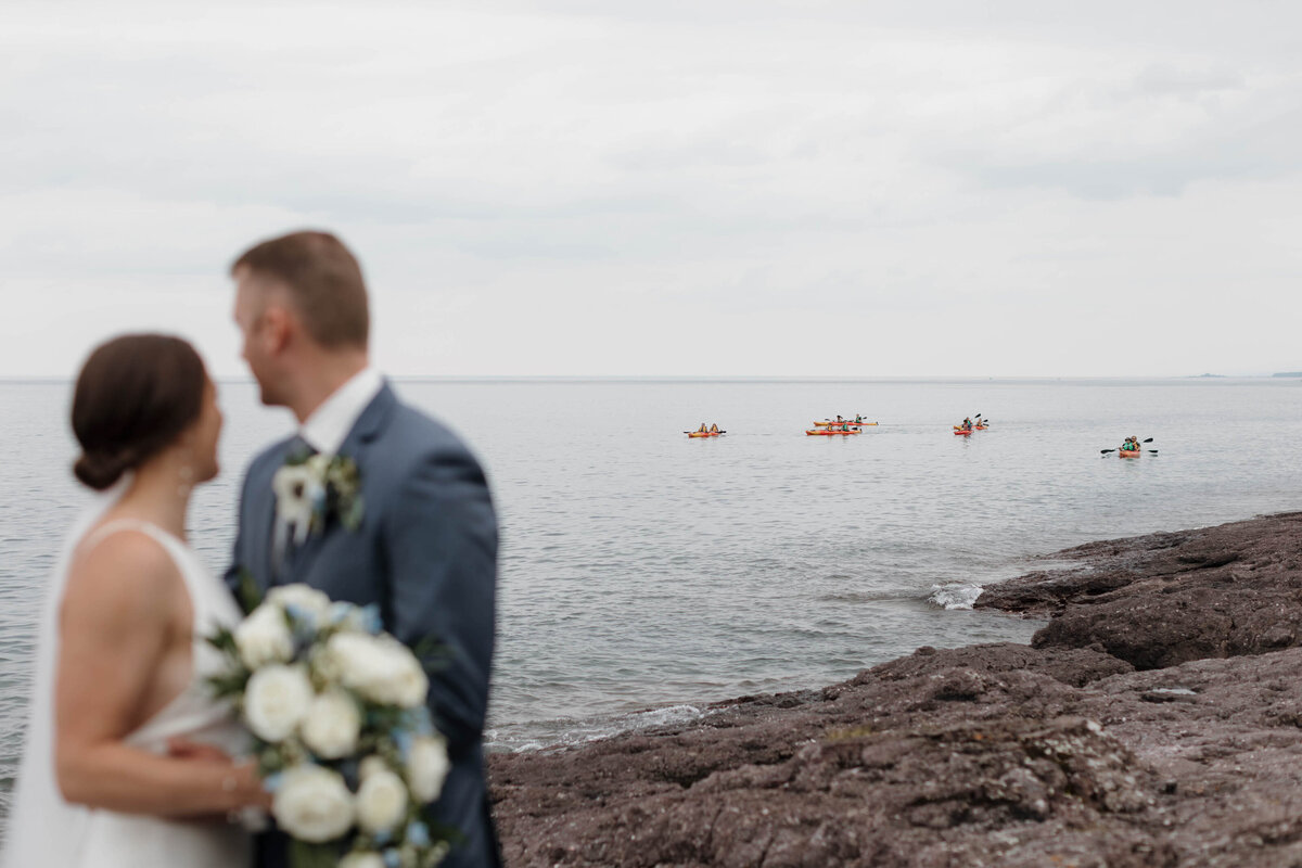 A newlywed couple standing at the water's edge looking out over it 