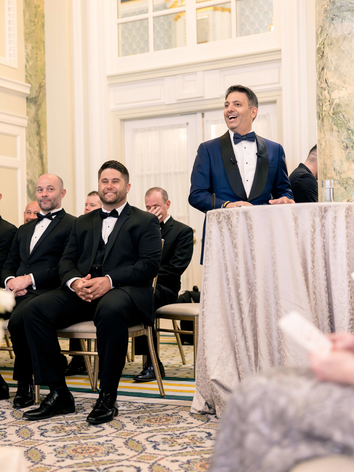 A man in a blue suit stands at a podium, speaking and smiling at a formal event. Seated nearby are two men in black tuxedos, listening attentively. The room has elegant décor with patterned carpet and tall windows.