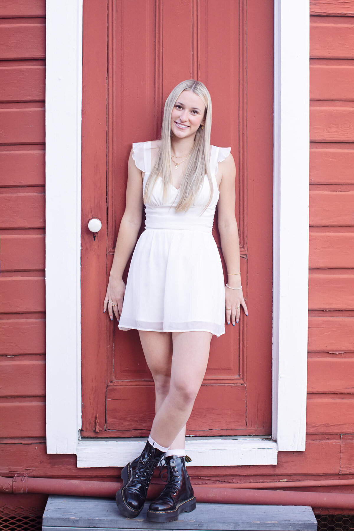 A woman with long blonde hair stands against a red wooden door. She is wearing a white dress and black boots. The door is part of a textured red wall. Her expression is relaxed and content.