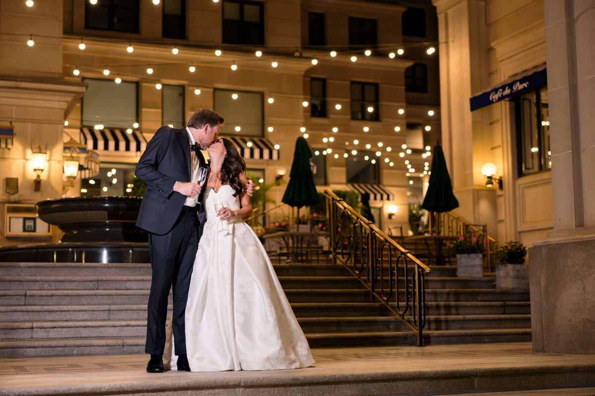 A bride and groom kiss in an elegant outdoor setting at night. They are standing on a stone staircase, surrounded by twinkling string lights and a large building with awnings in the background. The bride holds a champagne flute.