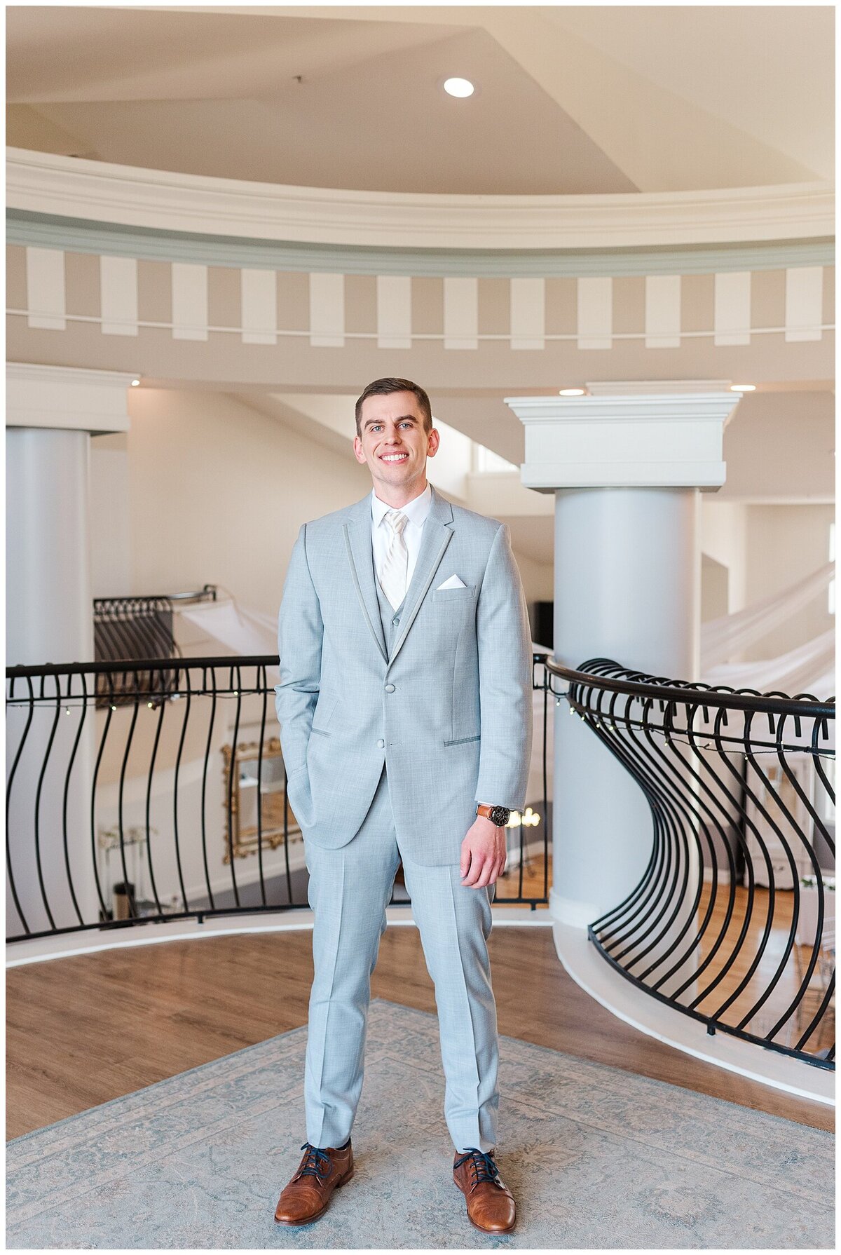 Groom in a light gray suit and brown shoes stands in the loft of the chateaux at fox meadows with one hand in his pocket as her smiles for denver wedding photographer