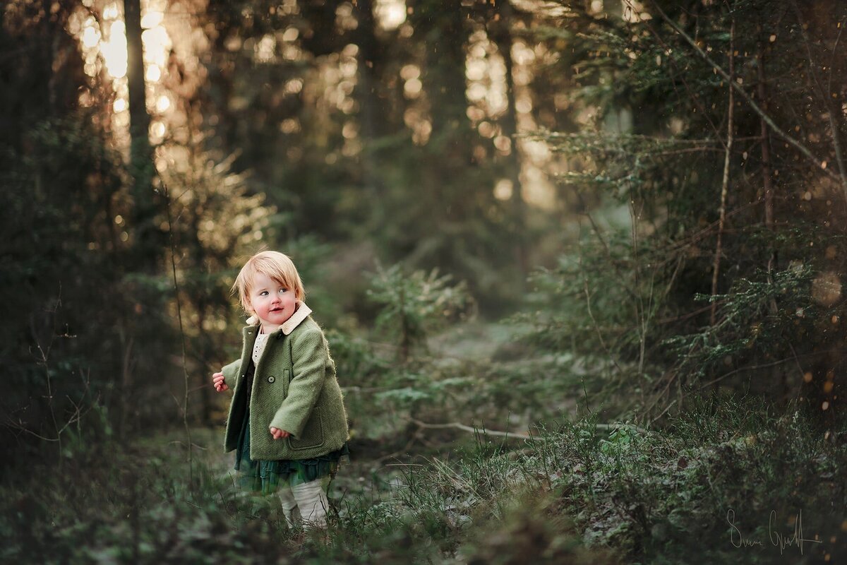 två-årig flicka med grön kjol från Newbie och en grön kappa står i skogen i veckert morgonljus och ser ser nyfiket omkring.