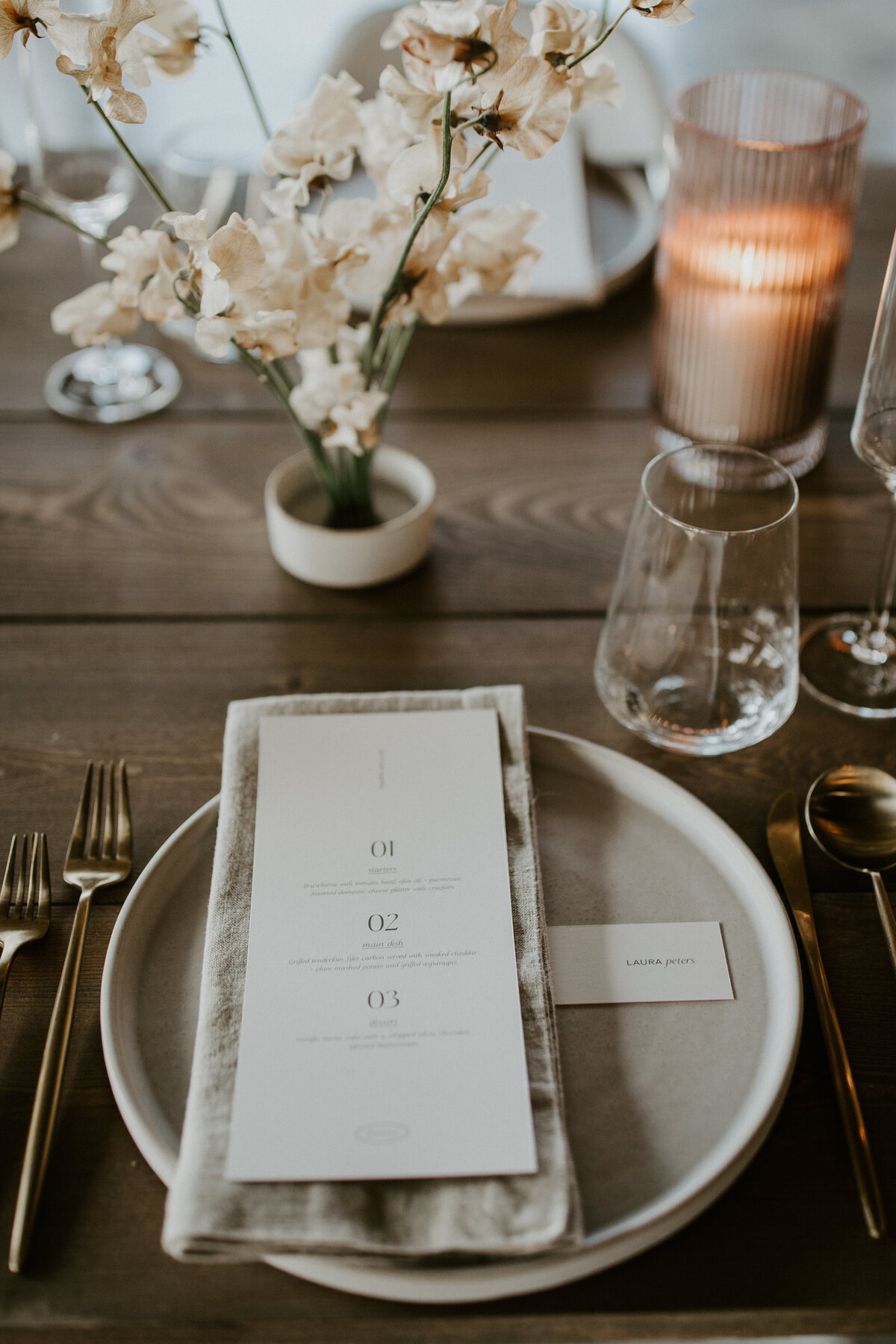 White dinner menu with black font atop a white linen napkin and white plate set on wooden table with white flowers.