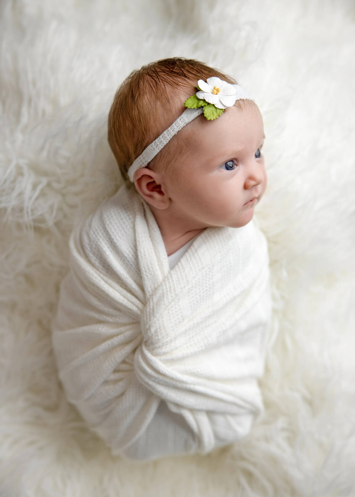 side profile of awake newborn baby wrapped in white fabric wearing a floral headband laying on a white shaggy rug