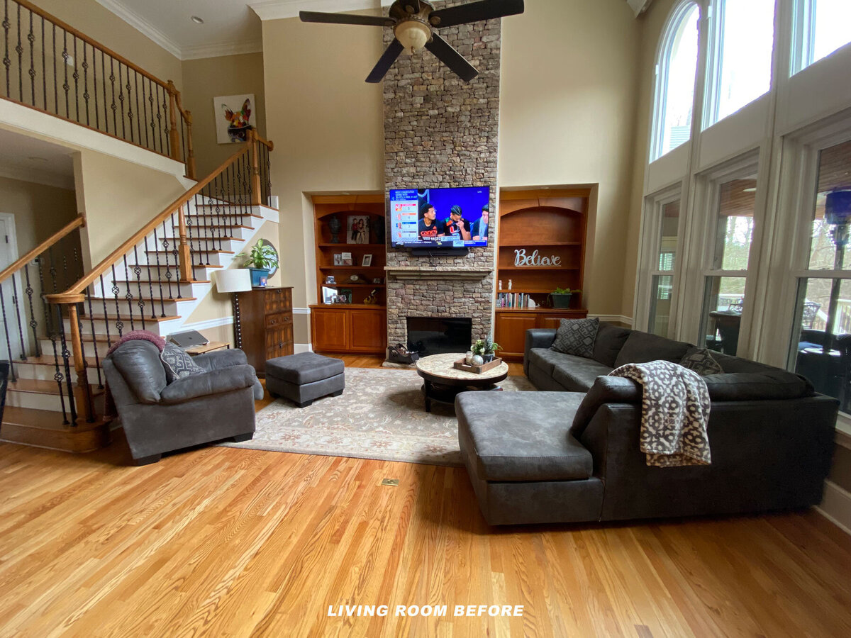 A spacious living room with wooden floors, large windows, and a stone fireplace. It includes a gray sectional sofa, an ottoman, a round wooden coffee table, and a flat-screen TV above the fireplace. "Living Room Before" is written on the floor.